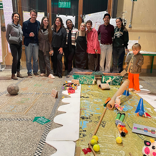 En grupp människor, framför dem ett barn som står i vad som ser ut som ett workshoparbete på golvet. Foto.