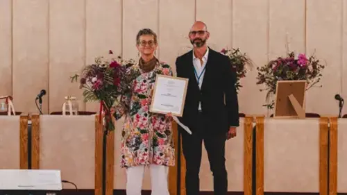 Maria Kylin, med diplom och blomsterbukett, och en man på scenen. Foto.