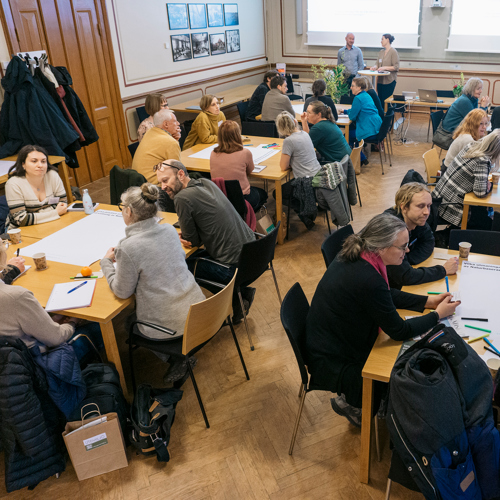 Människor sitter i grupper runt bord och diskuterar. Foto.