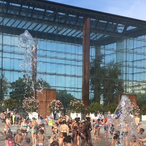Massor av barn svalkar sig i fontäner som sprutar upp ur marken på stort torg. Foto.