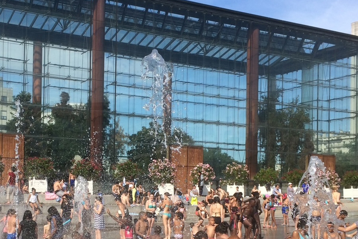 Massor av barn svalkar sig i fontäner som sprutar upp ur marken på stort torg. Foto.